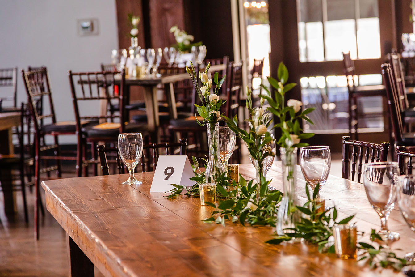 Corporate event tablescape with soft greenery, small white flowers, bud vases, and gold votive candles