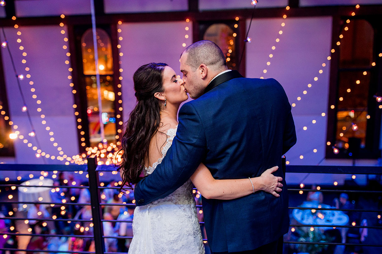 Bride and groom kiss on mezzanine overlooking wedding reception below