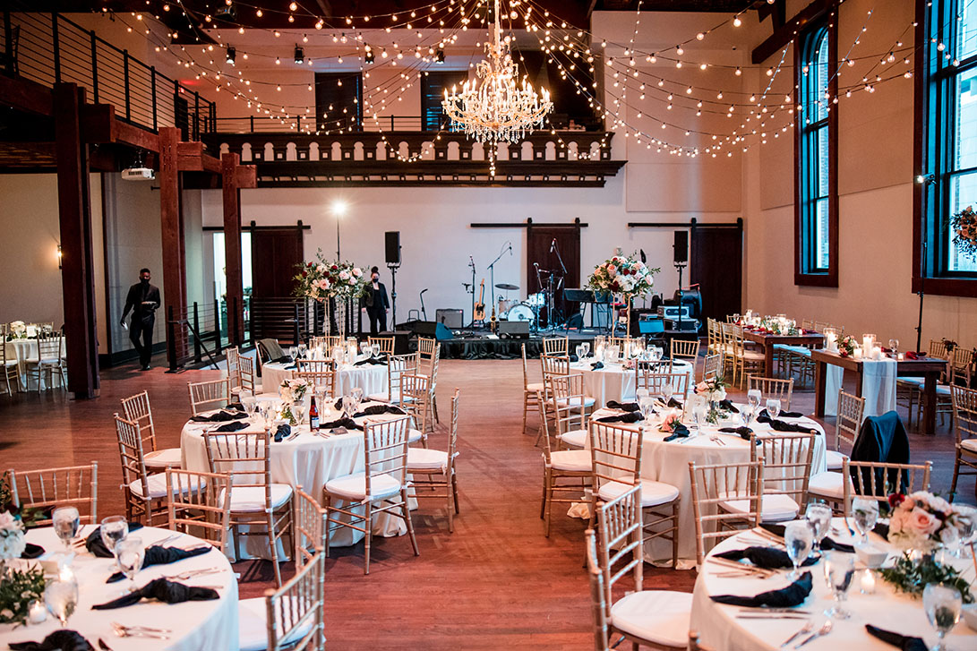 Wedding reception setup with round tables, chandelier, cascading string lights, and live music stage
