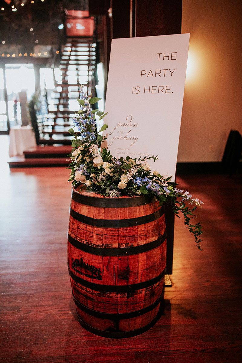 Wedding reception welcome sign on top of rustic barrel with abundant arrangement of blue flowers, white flowers, and greenery