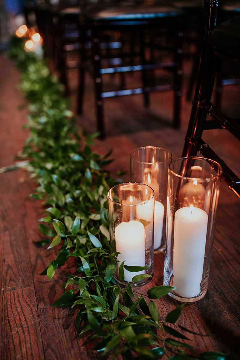 Greenery-lined aisle with white pillar candles in hurricane vases