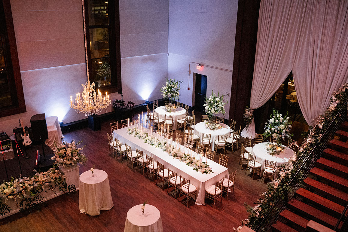Wedding reception setup with chandeliers, romantic tall arrangements, and soft pink uplighting