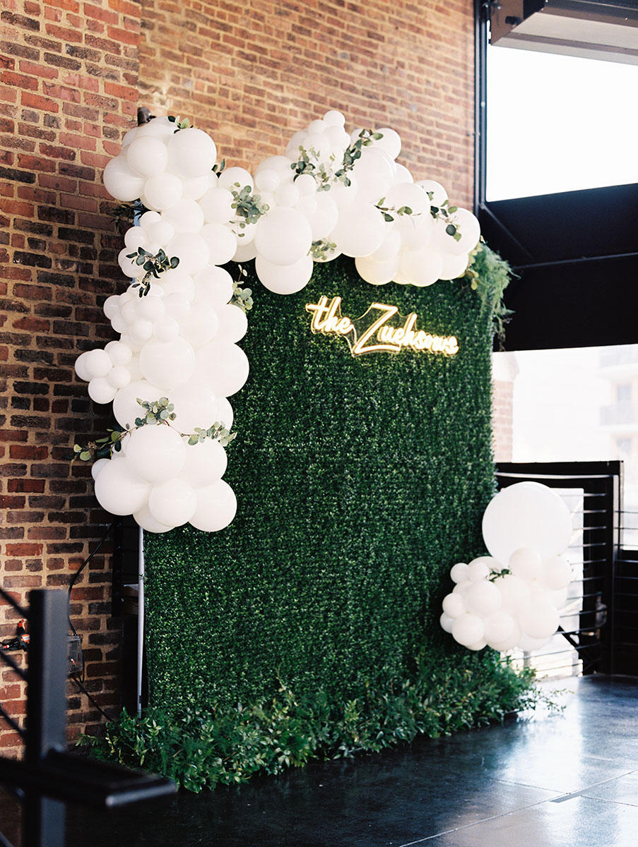 Photo wall backdrop setup with boxwood greenery, white balloon arch, and custom neon sign
