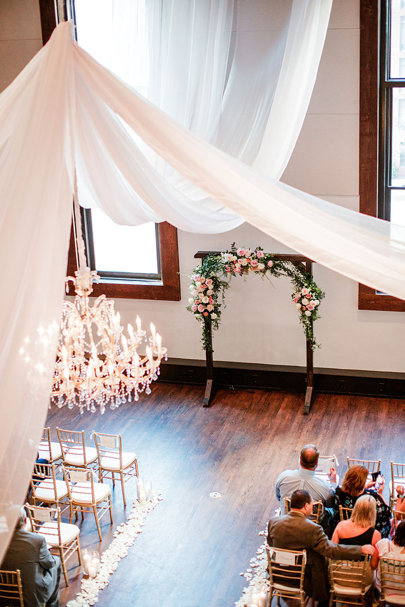 Wedding ceremony setup with drapery hanging from ceiling, wooden arch adorned with greenery and flowers, and a chandelier