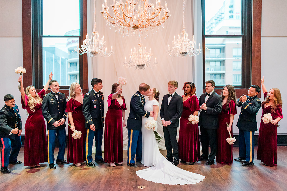 Bride and groom kiss under several chandeliers with cheering wedding party
