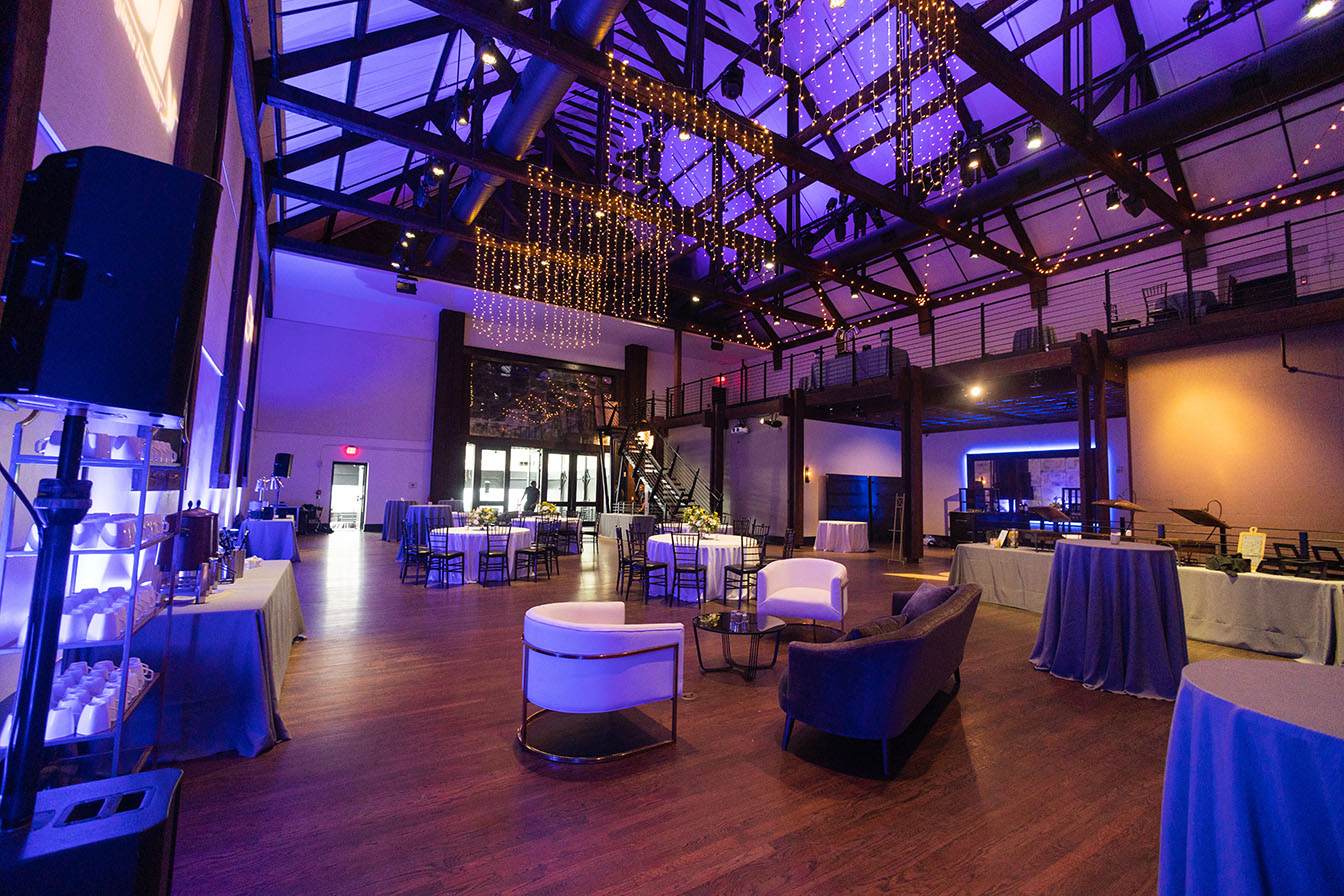 Corporate event setup in the main space with lounge area seating, round tables, string lights, and purple color wash