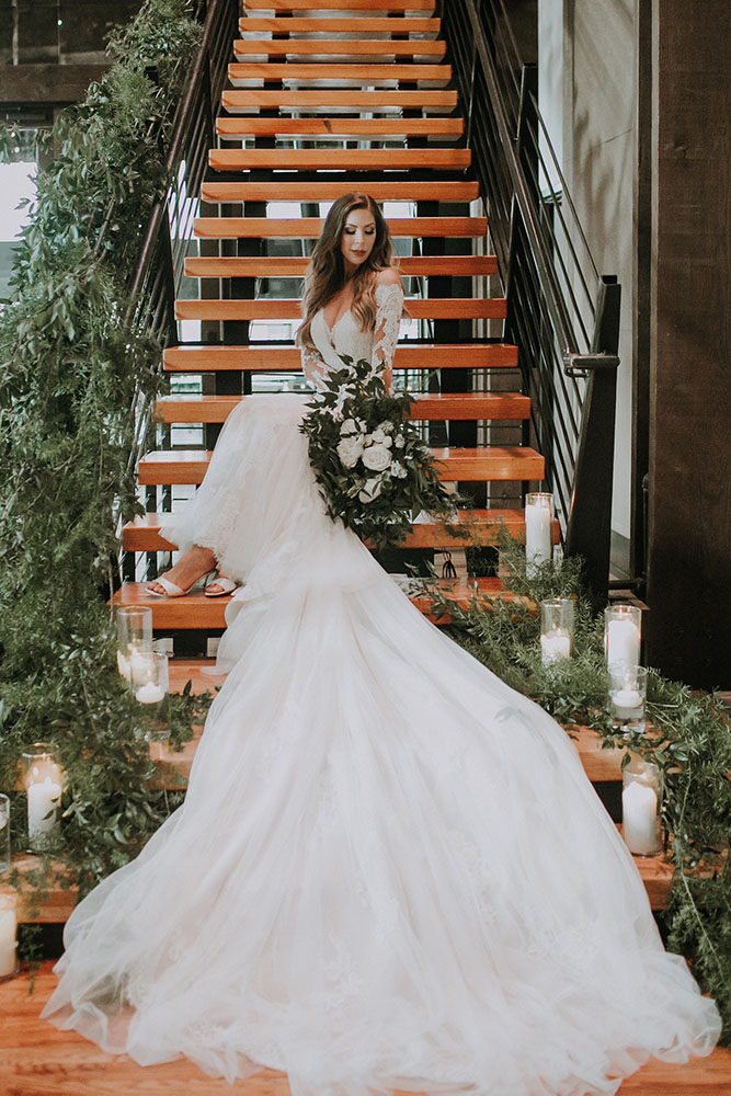 Bride sitting on staircase decorated in greenery wearing a long sleeve lace wedding gown.
