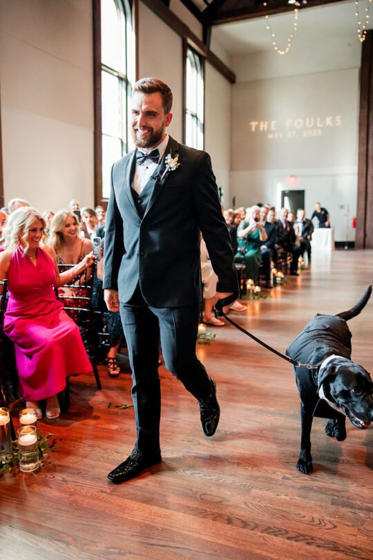 Groom walking down the aisle with a black lab