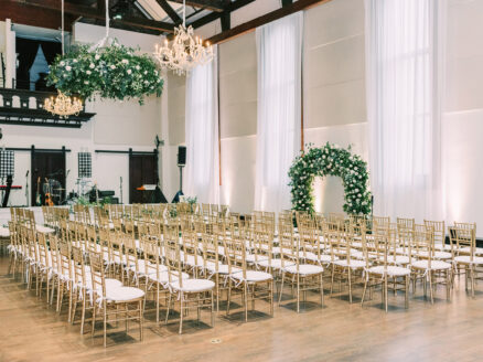 indoor wedding ceremony with greenery arch, hanging installation, and gold Chiavari chairs
